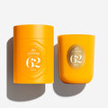 Two bright yellow-orange candles from Sol Janeiro Círculo 62 shown on a clean white background, one in a cylindrical tin and one in a rounded glass vessel, both with matching circular labels.
