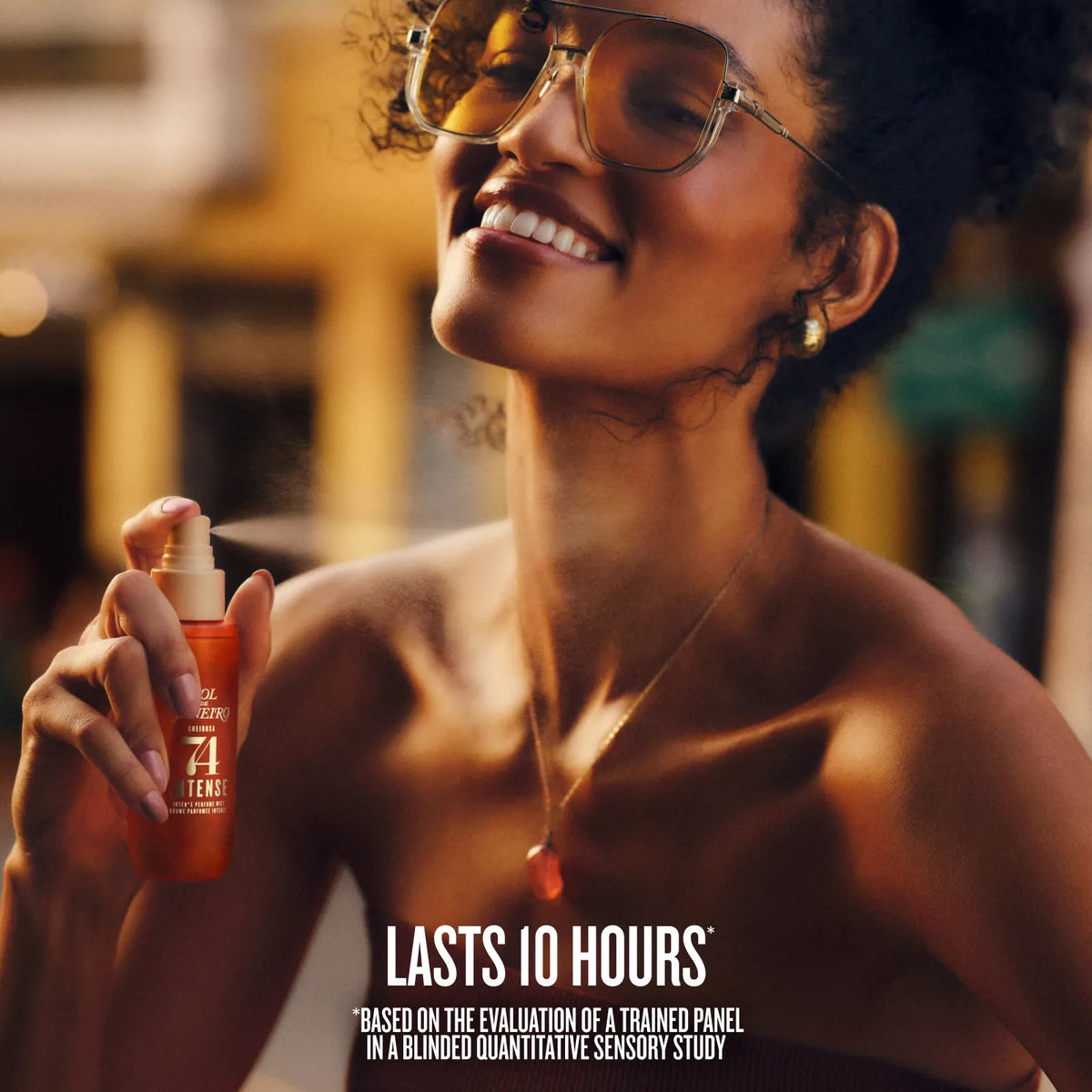 A woman wearing large tinted sunglasses and a gold necklace sprays a bottle of orange sunscreen on her neck in an outdoor setting. Text at the bottom reads 'LASTS 10 HOURS'.