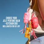 A close-up lifestyle shot of a person carrying a cream-colored shoulder bag with three colorful jelly perfume balm keychain bags attached. The bags are pink, coral red, and yellow, each with a small round bottle, keychain, and dangling charms.