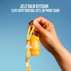 A hand holding a yellow and orange 'G2' jelly balm keychain with a yellow cord and silver carabiner, set against a clear blue sky. White text at the top reads 'JELLY BALM KEYCHAIN CLIPS ONTO YOUR BAG, KEYS, OR PHONE CHAIN'.