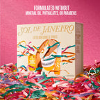 A boxed skincare gift set labeled SOL DE JANEIRO stands upright on a bed of colorful paper streamers against a pink background. The box shows illustrated bottles and jars with gold, teal, and peach accents.